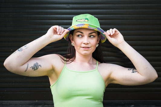 Woman wearing a green tank top and green corduroy bucket hat with colourful pattern on alternate side, against a dark background