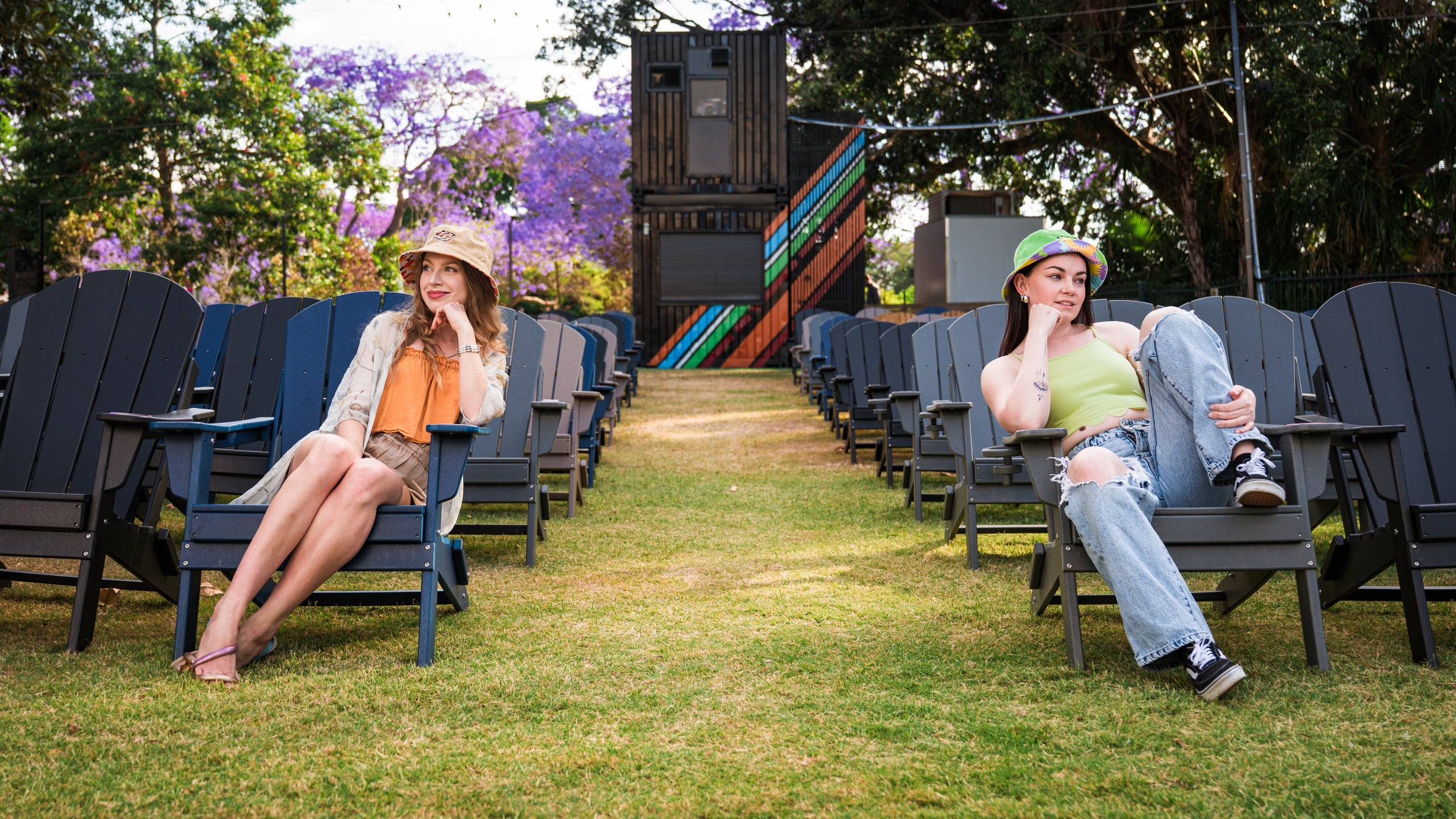 Two women wearing colourful bucket hats, sitting on chairs in a grassy area with trees and colorful striped structure in the background.