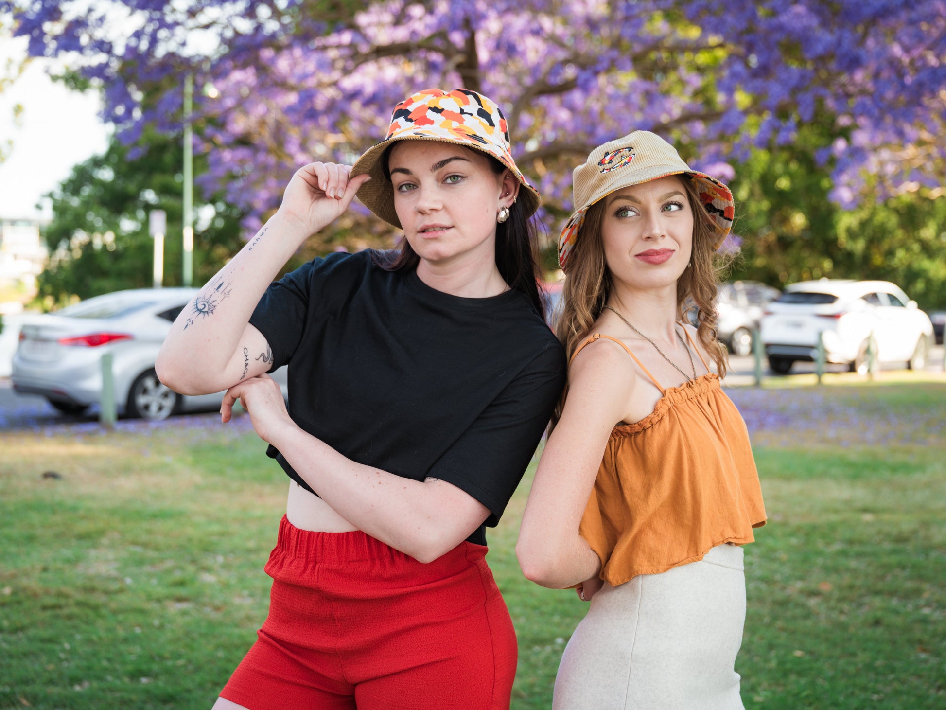 Two women wearing colourful koi fish bucket hats posing outdoors with a background of purple flowers and cars.