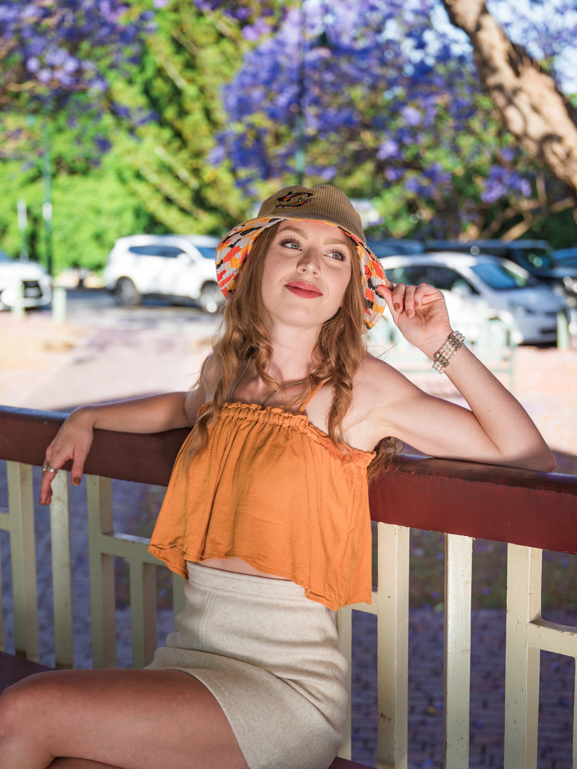 Woman in an orange top and beige skirt  with koi print bucket hat sitting on a railing with a blurred background